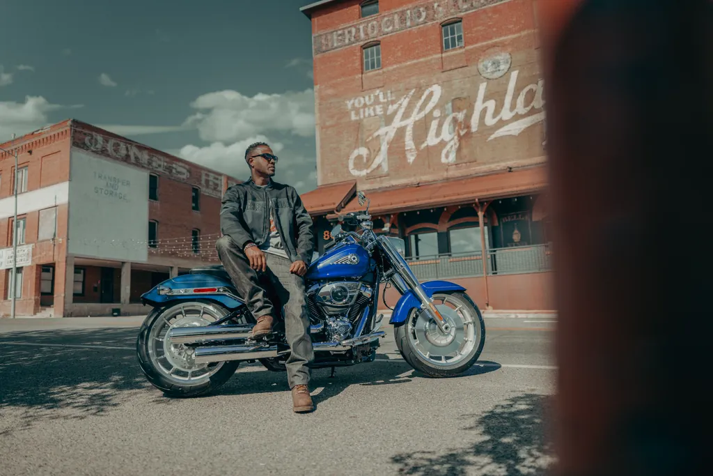 Une Harley-Davidson Fat Boy bleue de 2025, vue de profil dans une rue, avec son pilote appuyé sur la moto